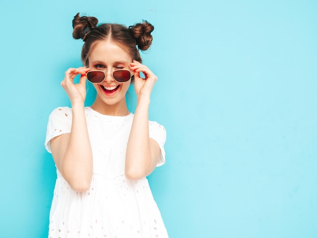 Portrait of young beautiful smiling female in trendy summer white dress carefree woman posing near blue wall in studio Positive model having fun indoors Cheerful and happy Takes off sunglasses