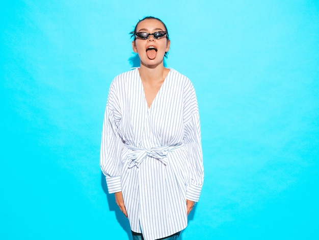 Free photo portrait of young beautiful smiling hipster girl in trendy summer clothes. sexy carefree woman posing near blue wall. positive model having fun in sunglasses.shows tongue