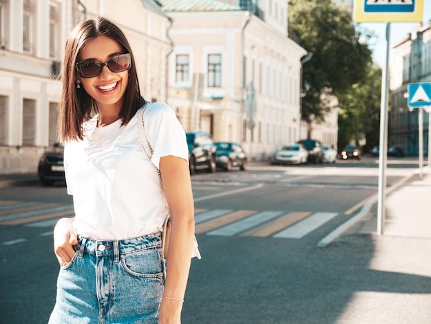 Portrait of young beautiful smiling hipster woman in trendy summer jeans shorts Sexy carefree model posing on the street background at sunset Positive model outdoors in sunglasses