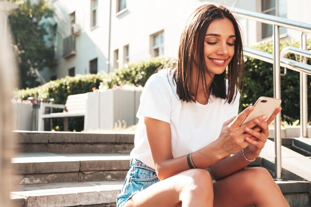 Portrait of young beautiful smiling hipster woman in trendy summer jeans shorts Sexy carefree model posing in the street Sitting at the stairsLooking at smartphone screen using phone apps