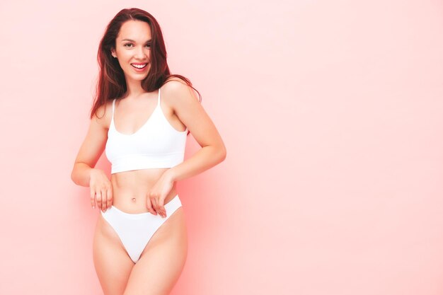 Portrait of young beautiful smiling woman in white lingerie. Sexy carefree cheerful model in underwear posing near pink wall in studio. Positive and happy female enjoying morning