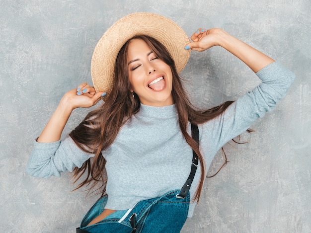 Portrait of Young beautiful smiling woman with closed eyes. Trendy girl in casual summer overalls clothes and hat. and shows tongue