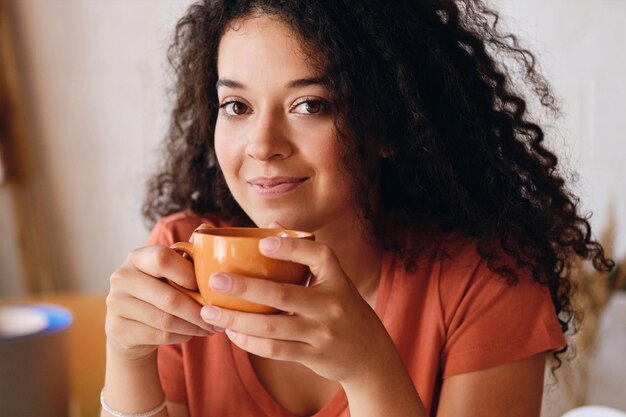 Portrait of young beautiful smiling woman with dark curly hair holding orange cup of coffee in hands dreamily looking in camera at modern cozy home alone