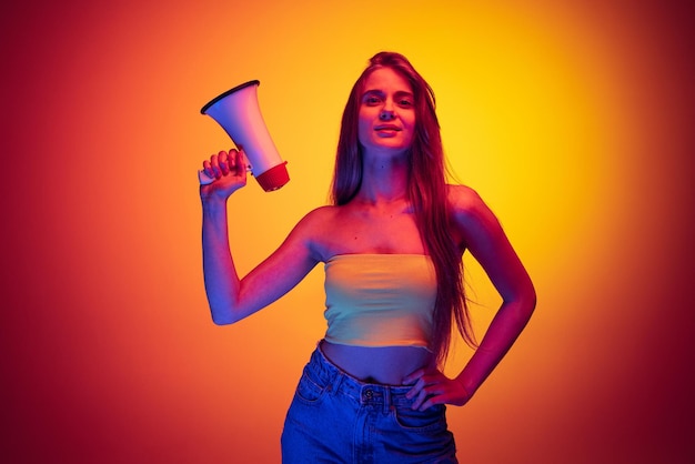 Portrait of young beautiful woman holding megaphone isolated over red yellow background in neon light