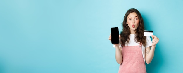 Free photo portrait of young beautiful woman showing plastic credit card and empty smartphone screen look amuse