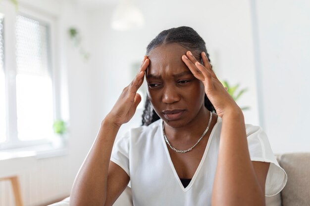 Portrait of a young black girl sitting on the couch at home with a headache and back pain Beautiful woman suffering from chronic daily headaches Sad woman holding her head because sinus pain