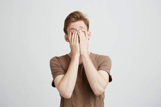 Portrait of young blond man closing face with hands.