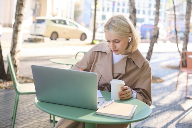 Free photo portrait of young blond woman with laptop sitting on street drinking coffee in outdoor cafe working