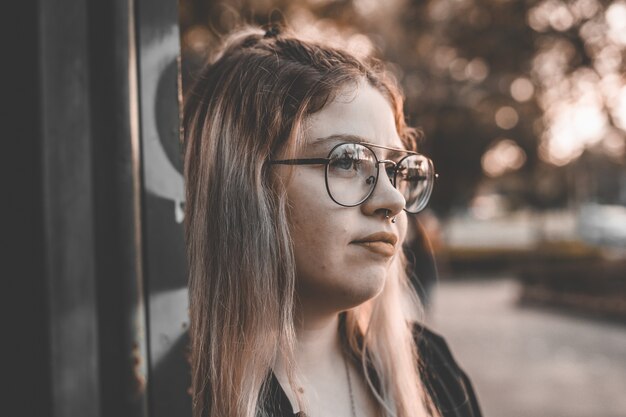 Portrait of a young blonde female with glasses leaning on a wall and smiling gently