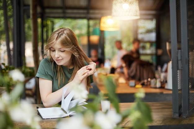 Portrait of a young blonde student woman reading her notes drinking coffee in a cafe outdoors waiting for her classes smiling.