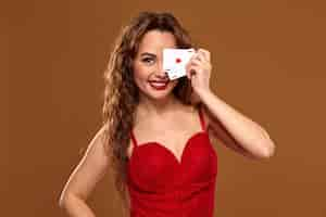 Free photo portrait of young or brown-haired woman smiling, holding pair of aces covering one eye with cards, wearing red cocktail dress on brown background. casino concept, gambling industry