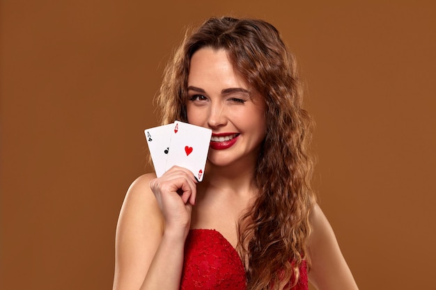 Free photo portrait of young or brown-haired woman smiling, holding pair of aces wearing red cocktail dress on brown background. casino concept, gambling industry