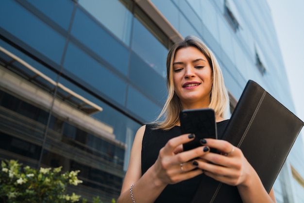 Portrait of young business woman using her mobile phone while standing outside office buildings. Business and success concept.