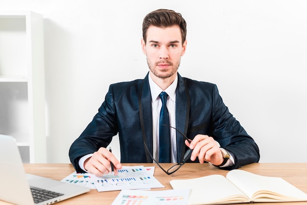 Free photo portrait of a young businessman holding pen over the graph and eyeglasses in hand looking to camera