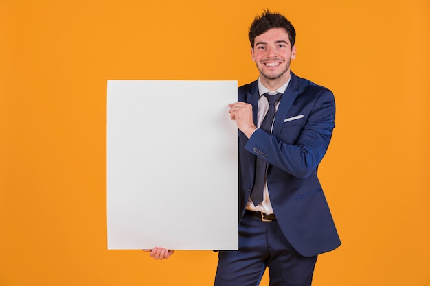 Free photo portrait of a young businessman holding white blank placard against an orange backdrop