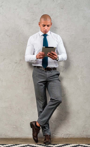 Free Photo portrait of a young businessman leaning on grey wall looking at digital tablet