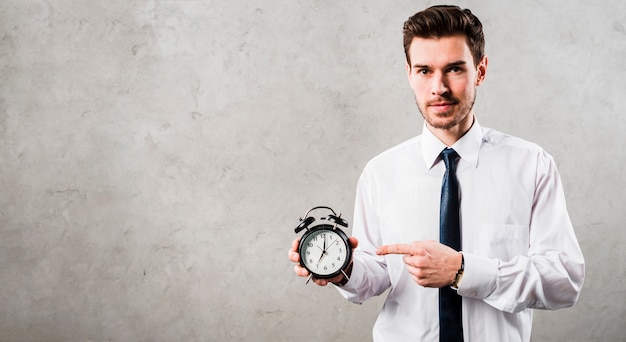 Free photo portrait of a young businessman pointing at black alarm clock standing against grey concrete wall