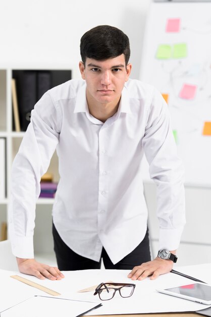 Portrait of a young businessman standing behind the desk looking at camera