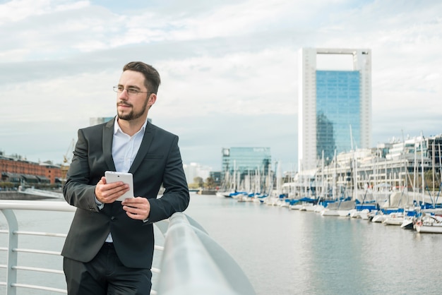 Portrait of a young businessman standing near the harbor holding mobile phone in hand looking away