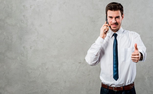 Portrait of a young businessman talking on mobile phone showing thumb up sign against concrete grey wall