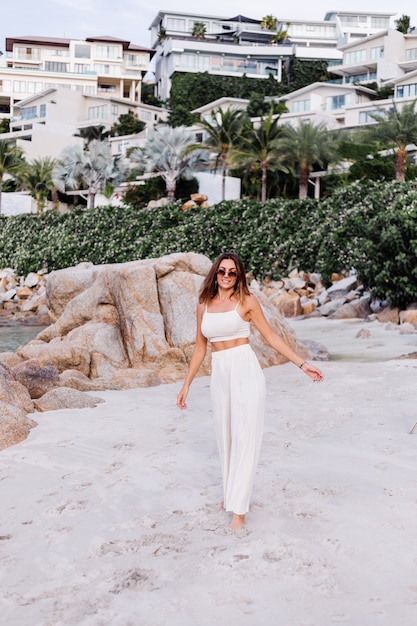 portrait of young calm happy caucasian fit slim woman in crop cami top and pants set alone on rocky tropical beach at sunset