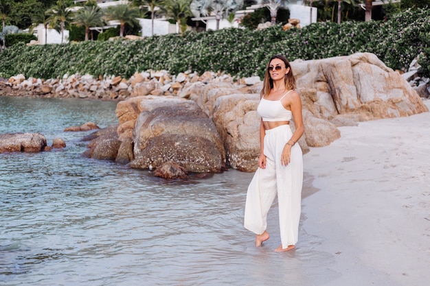 Free photo portrait of young calm happy caucasian fit slim woman in crop cami top and pants set alone on rocky tropical beach at sunset