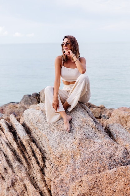 Free Photo portrait of young calm happy caucasian fit slim woman in crop cami top and pants set sits alone on rocky tropical beach at sunset