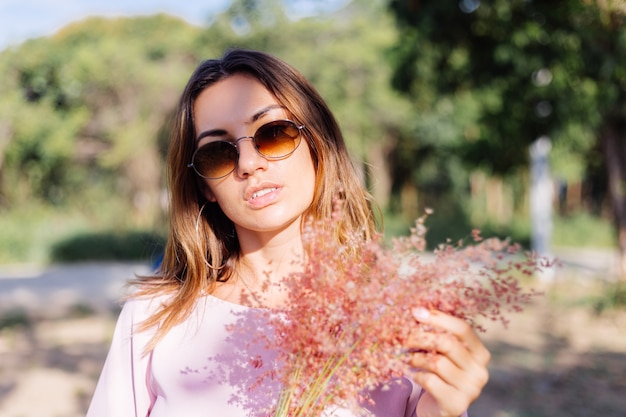 Portrait of young caucasian tanned woman in romantic pink dress round earings silver bracelet and sunglasses with wild flowers