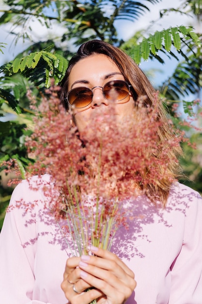 Free Photo portrait of young caucasian tanned woman in romantic pink dress round earings silver bracelet and sunglasses with wild flowers