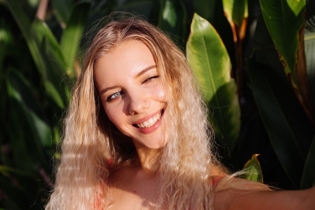 Portrait of young caucasian woman in red fitting elegant dress outdoor on background of tropical leafs