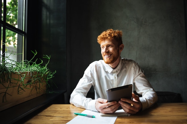 Portrait of young cheerful ginger bearded man, sitting at wooden table, holding tablet, looking at window