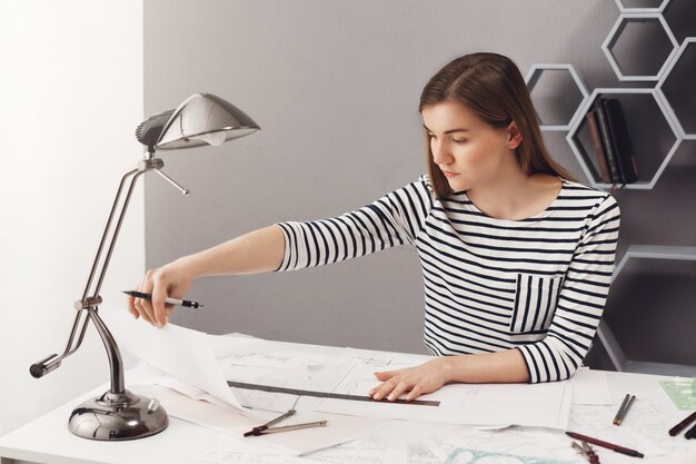 Portrait of young dark-haired student girl with long hair in striped shirt sitting at table in home, making architect project for exams, looking at drawings with concentrated face expression.