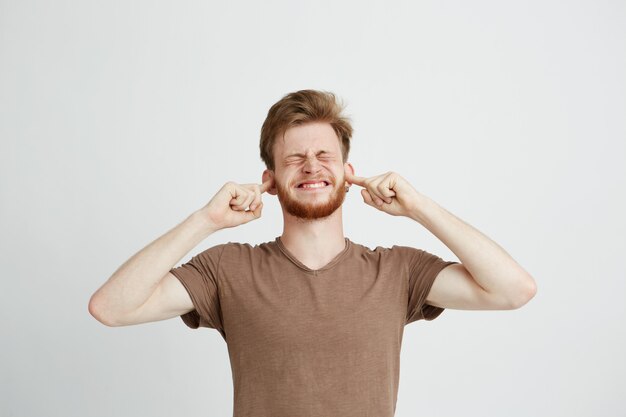 Portrait of young displeased angry man with beard closing ears.