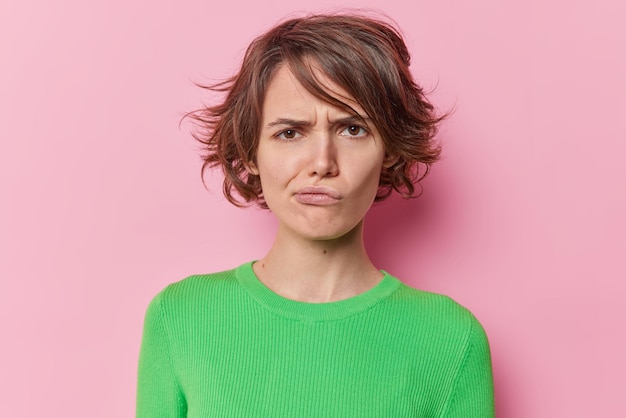 Free photo portrait of young displeased woman frowns face has sulking expression purses lips has short hairstyle wears green jumper isolated dover pink studio background. negative human emotions concept