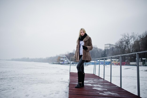 Portrait of young elegance blonde girl in a fur coat at pier background foggy river on winter ice