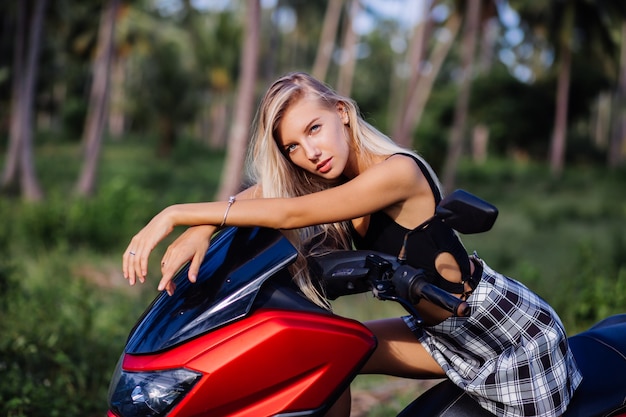 Free photo portrait of young european caucasian fit slim woman in black top swimsuit and plaid black and white school girl skirt on tropical jungle field with motorbike, motorcycle. sunglasses and black cap