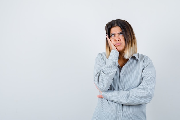 Portrait of young female leaning cheek on hand in oversized shirt and looking pensive front view