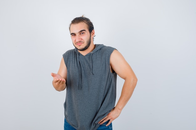 Portrait of young fit male spreading palm at camera in sleeveless hoodie  and looking hesitant front view