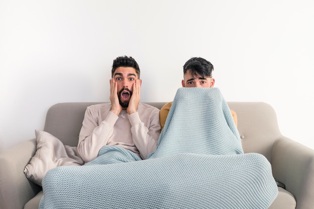 Portrait of young gay couple sitting on sofa watching horror movie on television against white wall