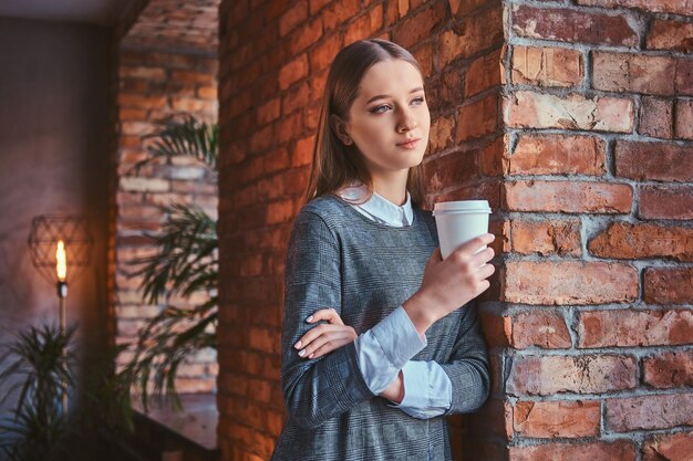 Portrait of a young girl dressed in an elegant gray dress leaning against a brick wall holds a cup of takeaway coffee looking away.