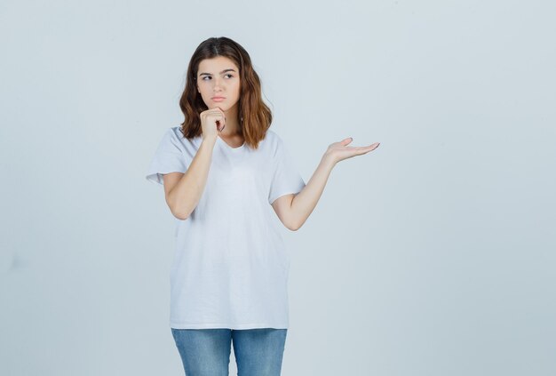 Portrait of young girl propping chin on hand, spreading palm aside in white t-shirt and looking pensive front view