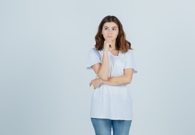 Portrait of young girl propping chin on hand in white t-shirt and looking pensive front view