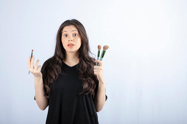 Portrait of young girl with cosmetic brushes looking at camera on white.