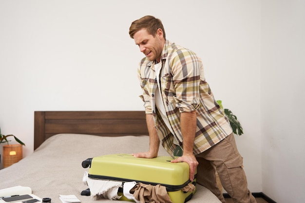 Free photo portrait of young guy traveller going on holiday pressing his suitcase trying to push clothes inside