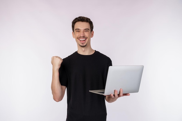 Portrait of young handsome smiling man holding laptop in hands typing and browsing web pages while doing a winning closed fist gesture isolated on white background