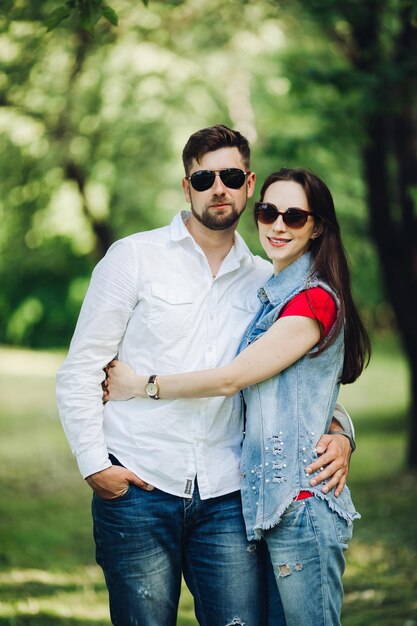 Portrait of young happy couple in love smiling and embracing in garden Sweet lovers wearing in stylish dark sunglasses and casual clothes posing and looking at camera at daytime in summer park