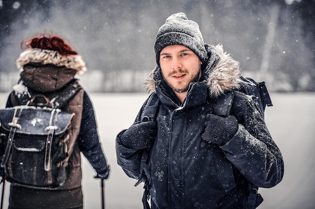 Portrait of a young hiker guy with a backpack walking with his girlfriend through a winter forest