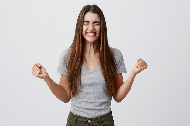 Portrait of young joyful happy beautiful female student with long dark hair in casual gray outfit smiling with teeth, spreading hands with clothed eyes, being extremely happy finally get role in movie