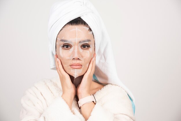 Portrait of young lady in bathrobe and towel on head while touching her face with facemask