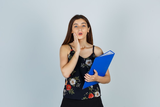 Portrait of young lady holding folder, keeping hand on cheek in blouse, skirt and looking wondered front view